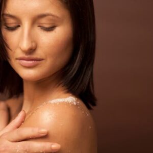 Close-up of a woman enjoying a skincare routine, with exfoliating salt on her shoulder, in a warm studio setting.