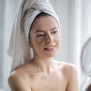 A smiling woman wrapped in a towel looks into a handheld mirror indoors, enjoying her skincare routine.