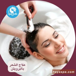 امرأة تبتسم وعيناها مغمضتان أثناء غسل شعرها بعد تنعيم الشعر بالبروتين والكيراتين في صالون تجميل، ويد مصفف شعر تمسك برأس الدش لصب الماء على شعرها.