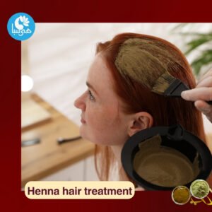 A young woman with red hair sits on a chair as a hairdresser applies a henna mixture to her scalp and hair using a brush