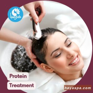 A woman smiling with her eyes closed as her hair is being washed after did hair protein and keratin treatment in a salon, with a hairdresser's hand holding a shower head to pour water over her hair.