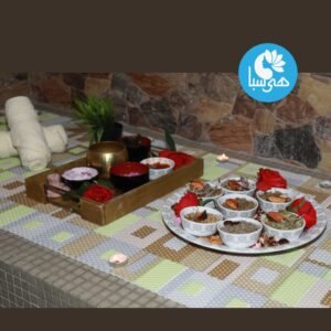 صينية وطبق مرتبان بشكل جميل مع مكونات طبيعية متنوعة لخدمة الحمام المغربي في هي سبا. تتضمن العناصر أوعية من المقشرات والزيوت والأعشاب، مزينة بالورود الحمراء والشموع، على خلفية من المناشف وجدار مزخرف. شعار هي سبا ظاهر في الزاوية العلوية اليمنى.
